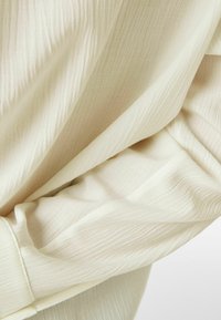 Jasnobeżowa tkanina z teksturowanym, falistym wzorem, delikatnie opadająca i ukazująca subtelne fałdy. Materiał wydaje się lekki i gładki.