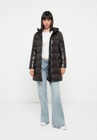 Sort puffet jakke med en blank overflade, der har en lynlås foran, store quiltede sektioner og en hætte, parret med lyseblå flagrende jeans.