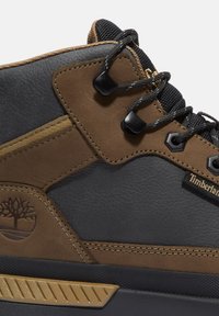 Zapatilla superior de cuero negro y marrón con acabado texturizado, acentos dorados, logotipo prominente y cordones negros asegurados por ojales negros.