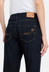 I jeans in denim scuro presentano un alto punto vita, cuciture gialle a contrasto e due tasche posteriori con un logo stilizzato e un design curvo.
