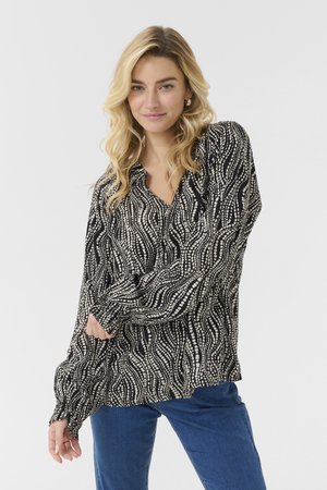Femme blonde aux cheveux ondulés portant un chemisier à manches longues à motifs noir et blanc et un jean bleu, souriant les bras croisés.