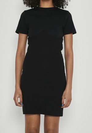 Vrouw die een nauwsluitende, zwarte jurk met korte mouwen tot op de knie draagt, staand tegen een lichtgrijze achtergrond.