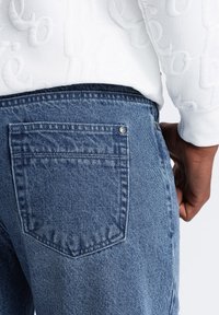 Jeans de mezclilla azul con una textura suave, que cuentan con un bolsillo trasero y un detalle de botón de metal. Acompañados de un suéter blanco texturizado.