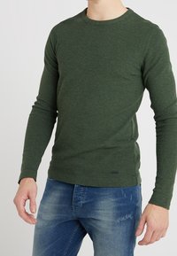 Mörkgrön långärmad tröja med rund halsringning, tillverkad av ett texturerat tyg, parat med ljusblå denimjeans med en slitna mönster.