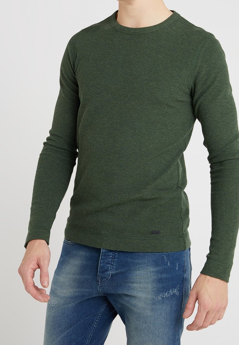 Mörkgrön långärmad tröja med rund halsringning, tillverkad av ett texturerat tyg, parat med ljusblå denimjeans med en slitna mönster.