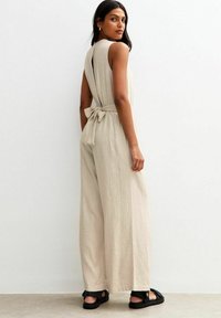 Combinaison en lin beige avec un détail de nœud dans le dos, silhouette à jambes larges et design sans manches. Associée à des sandales noires.
