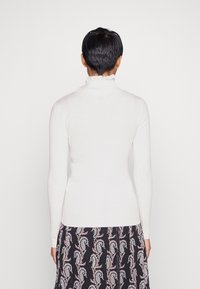 Weißer gerippter Rollkragenpullover mit einem Rüschensaum und taillierter Silhouette. Kombiniert mit einem gemusterten schwarzen Rock mit Paisley-Muster.