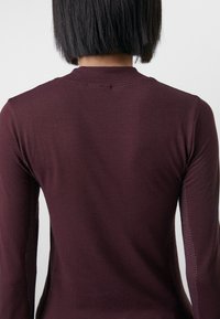 Mörkröd långärmad topp i texturerat material, med mesh paneler på sidorna och rund halsringning. Slät yta utan synlig hårdvara.