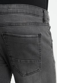 Jeans de mezclilla gris con un bolsillo trasero de forma rectangular y costuras en contraste, mostrados desde un ángulo lateral.
