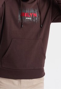 Brązowa bluza z kapturem z przednią kieszenią. Na froncie znajduje się grafika z napisem "BKLYN" na czerwono oraz "NEW YORK CITY" na biało. Bluza posiada sznurki i ściągacze na rękawach.