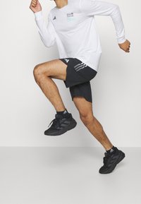 Chemise blanche à manches longues avec logo imprimé, associée à un short noir avec une bande latérale. Des chaussures de sport noires complètent la tenue.