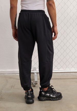 Osoba oblečená v černých teplákových kalhotách a černých teniskách s bílým logem, stojící na betonové podlaze před pozadím z drátěné sítě.