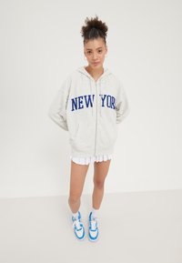 Sweat à fermeture éclair gris clair avec "NEW YORK" en lettres bleues, associé à une jupe plissée blanche et des baskets avec des accents bleus. Texture douce.