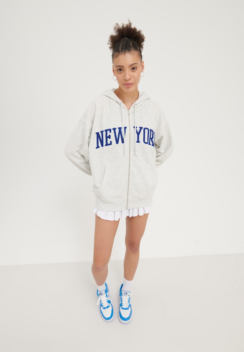 Sweat à fermeture éclair gris clair avec "NEW YORK" en lettres bleues, associé à une jupe plissée blanche et des baskets avec des accents bleus. Texture douce.