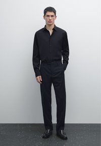 Czarna koszula z guzikami i kołnierzykiem, w połączeniu z ciemnymi spodniami i wypolerowanymi butami. Stylizacja charakteryzuje się eleganckim, minimalistycznym designem.