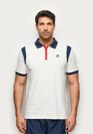 Hombre con pelo corto y oscuro lleva una camisa polo blanca con cuello y mangas azul marino, tapeta de botones roja y pantalones cortos azul marino, de pie sobre fondo liso.