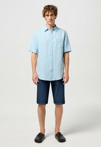 Chemise à manches courtes bleu clair avec une poche poitrine, associée à un short bermuda en denim foncé et des sandales noires à enfiler.