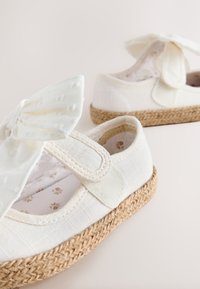 White fabric baby shoes with woven jute soles, Velcro straps, and decorative bows on a light background.