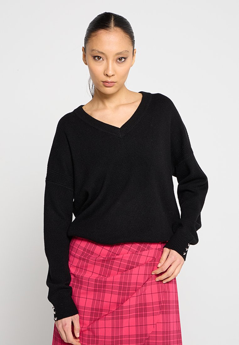 Maglione nero lavorato a maglia con scollo a V e vestibilità ampia. Le maniche lunghe presentano bottoni decorativi ai polsini. Indossato sopra una gonna a quadri rosa.