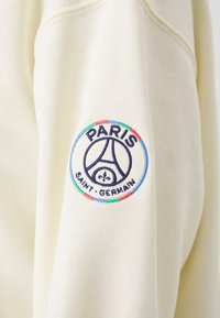 Logo del Paris Saint-Germain bordado en la manga de un sudadera color crema, que presenta la Torre Eiffel y el símbolo de la flor de lis.
