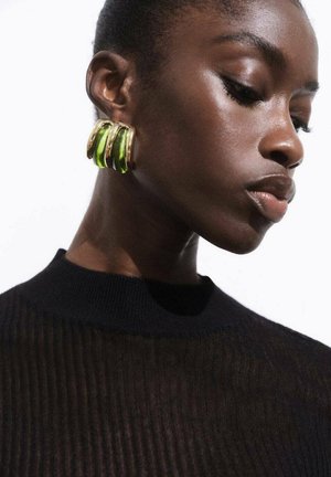 Gouden hoepel oorbellen met drie groene accenten, met een glanzende afwerking. Het model draagt een zwart, ribbelgebreid top.