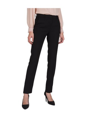 Kvinde iført sort slim-fit bukser, lyserød bluse med blonde manchetter og sorte højhælede sko, stående mod en hvid baggrund.