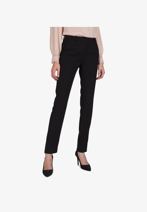 Woman wearing slim-fit black trousers, pale pink blouse with lace cuffs, and black high-heeled shoes, standing against white background.