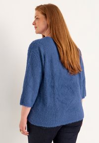 Blauer, flauschiger Pullover mit Dreiviertelärmeln, der einen lässigen Schnitt und eine strukturierte Oberfläche aufweist, kombiniert mit dunklen Jeans.