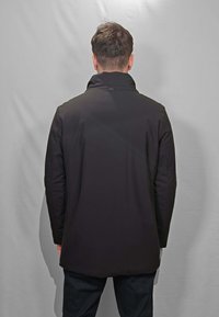Cappotto nero impermeabile con collo alto e maniche lunghe. Tessuto liscio, leggermente più lungo nella parte posteriore, con minimalista accessorio.