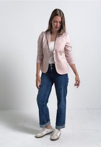 Blazer rose clair à un bouton et manches retroussées, associé à un jean bleu foncé à revers et des baskets blanches. Design simple et moderne.