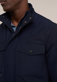 Man med en marinblå jacka med dragkedja och tryckknappsstängning samt framfickor över en ljusblå skjorta.