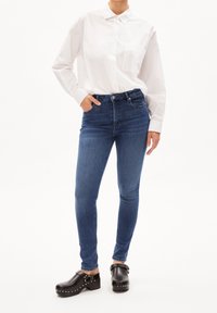 Tmavě modré skinny džíny spárované s bílou košilí s dlouhým rukávem. Černé dřeváky se cvočky. Hladká textilie a klasický design límce.