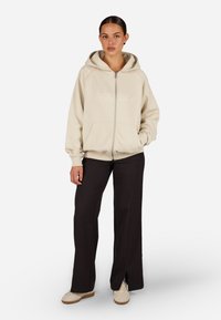 Beige zipzáras kapucnis pulóver kenguruzsebekkel, hímzett felirattal. Fekete, bővített nadrággal és világos bézs textúrájú cipővel párosítva.