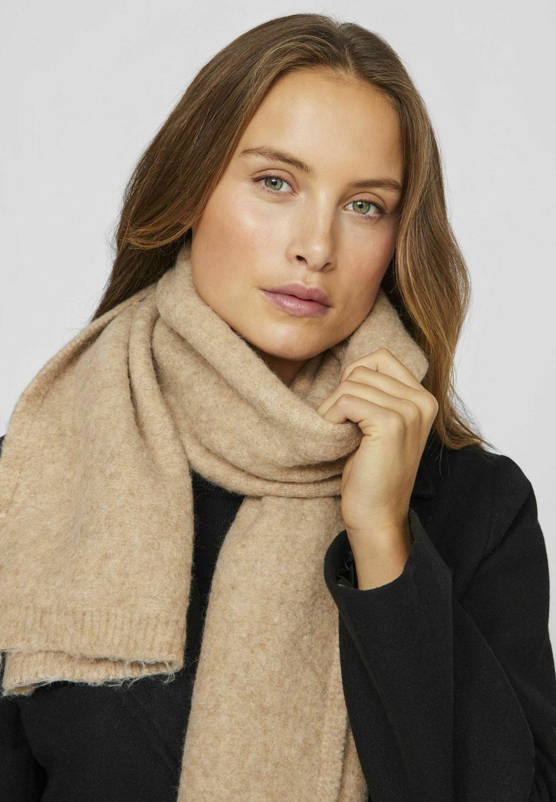 Beige wollen sjaal met een zachte textuur, voorzien van rafelige randen en een geribbelde rand. Om de nek gewikkeld, contrasterend met een zwarte jas.