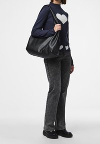 Sac hobo noir en matériau texturé, affichant une forme souple et décontractée. Porté à l'épaule, complété par un pull bleu marine avec des cœurs blancs.