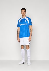 Man in een blauw-witte voetbaloutfit, met witte sokken en scheenbeschermers, en witte voetbalschoenen, staand tegen een effen lichte achtergrond.