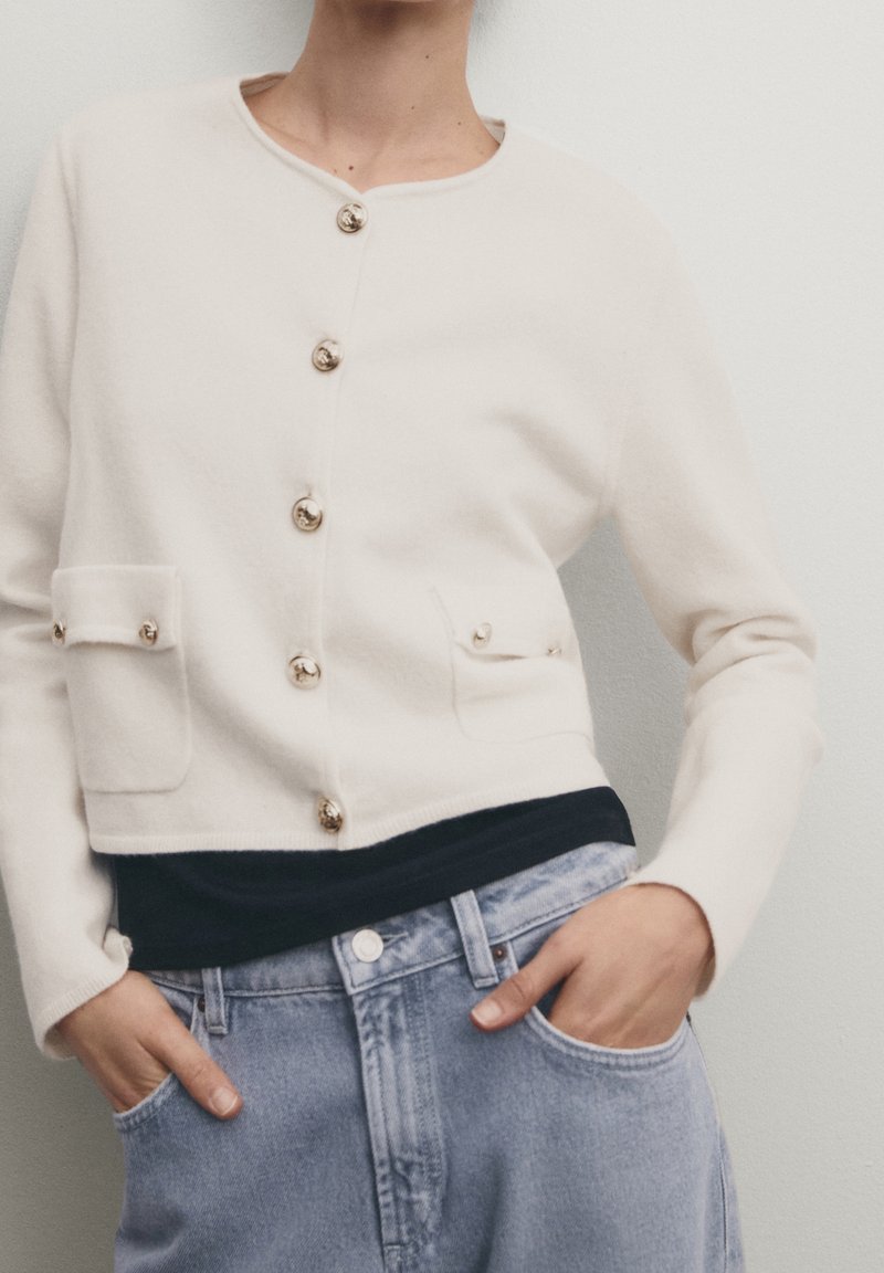 Cream cardigan with a round neckline, golden buttons, and two front pockets. Fabric appears soft and textured. Paired with a dark top and light jeans.