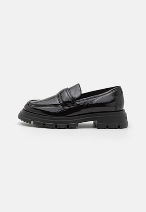 Mocassin en cuir verni noir avec un bout arrondi, semelle en caoutchouc épaisse et une couture minimale. Présente une petite sangle sur le devant.