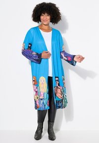 Blue lightweight cardigan with vibrant illustrated characters on sleeves, open front design, and a relaxed fit. Black fitted pants and studded boots.