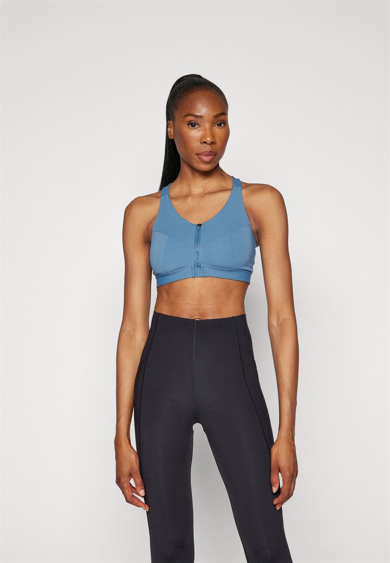 Μπλε αθλητικό σουτιέν με φερμουάρ μπροστά, φαρδιές τιράντες και εφαρμοστή γραμμή. Συνδυάζεται με μαύρες κολάν με ψηλή μέση. Λείο υφάδι, ελάχιστες ραφές.
