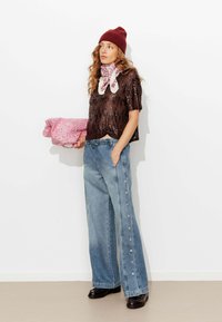 Haut marron pailleté, jean bleu clair à jambes larges avec des boutons sur le côté, sac à motifs roses, écharpe blanche et bonnet en tricot bordeaux. Chaussures noires.