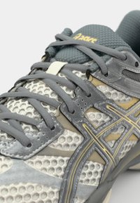 Gray athletic shoe with mesh upper, textured accents, yellow detailing, and gray laces. Features a padded tongue and breathable design.