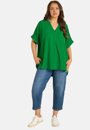 Vrouw draagt een los vallende groene top met V-hals, korte blauwe spijkerbroek en witte sneakers, staat met handen in de zakken tegen een effen achtergrond.