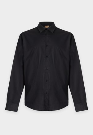Homme debout portant une chemise noire boutonnée, un pantalon marron, des chaussures noires, une main dans la poche, sur un fond clair uni.