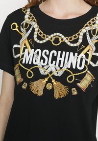 Svart bomullströja med en stor grafisk design som har guld saxar, sömnadsverktyg och "MOSCHINO" i fet stil med vita bokstäver.
