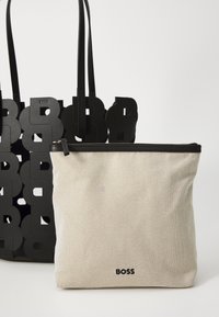 Bolso tote negro con diseño de logo recortado, acompañado de un neceser beis con cremallera que presenta una superficie texturizada y una marca en negro.