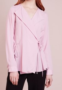 Vrouw in een lichtroze wikkelblouse met lange mouwen en zijknopen, gecombineerd met zwarte broek, tegen een neutrale achtergrond.