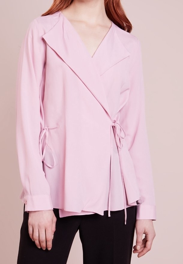Vrouw in een lichtroze wikkelblouse met lange mouwen en zijknopen, gecombineerd met zwarte broek, tegen een neutrale achtergrond.