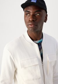 Chaqueta bomber blanca con cremallera y un patrón texturizado, combinada con una camisa de estampado multicolor. Gorro negro con logotipo.