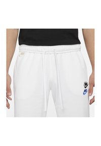 Vita Nike-joggers med elastisk midjeband, dragsko och två anmärkningsvärda logotyper: en svart och blå Nike-emblem. Slät, mjuk tygtextur.
